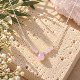 Rose quartz beaded necklace featuring a soft blush-pink teardrop pendant, delicate pink gemstone beads, and sterling silver chain, displayed on natural beige stone with olive branches, white flowers, and rose quartz crystal.