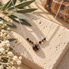 Tiger’s Eye beaded necklace featuring a faceted brown and golden teardrop pendant on a delicate silver chain, displayed on natural beige stone with olive branches and white flowers.