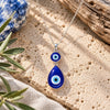 Large blue double evil eye necklace featuring a round and teardrop-shaped glass pendant on a silver chain, displayed on natural travertine stone with olive branches and soft daylight.