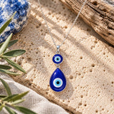 Large blue double evil eye necklace featuring a round and teardrop-shaped glass pendant on a silver chain, displayed on natural travertine stone with olive branches and soft daylight.