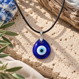 Blue glass evil eye necklace with black leather cord and silver-tone bail, displayed on natural travertine stone with olive branches and soft daylight.