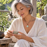 Woman wearing a delicate green cord necklace with a small carved jade Buddha pendant and red accent bead, styled with light-colored clothing in a serene outdoor setting.