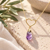 Gold open-heart necklace with a dangling raw purple amethyst crystal pendant, displayed on textured natural stone with soft greenery and ivory linen in natural daylight.