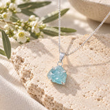 Raw aquamarine crystal necklace with a silver chain and prong-set pale blue gemstone pendant, displayed on textured natural stone with delicate white flowers.