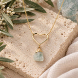 Raw aquamarine crystal necklace with a gold open-heart pendant and delicate chain, styled on natural limestone with olive branches and soft neutral accents.