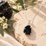 Raw black tourmaline crystal pendant in a silver prong setting on a delicate silver chain, displayed on textured beige stone with white flowers and natural greenery.