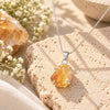 Raw golden citrine crystal pendant in a silver setting on a delicate chain, displayed on textured beige stone with white flowers and natural accents.
