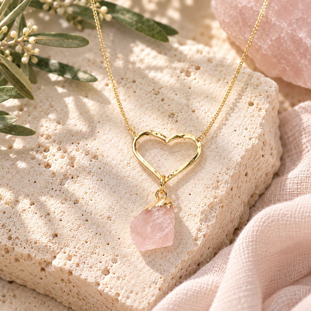 Raw Rose Quartz Heart Crystal Necklace with a delicate gold open-heart pendant and pale pink crystal, displayed on natural limestone with olive branches and soft blush linen.