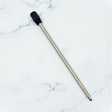A replacement pen refill with a long metallic silver body and a black textured cap, placed diagonally on a white marble-patterned background.
