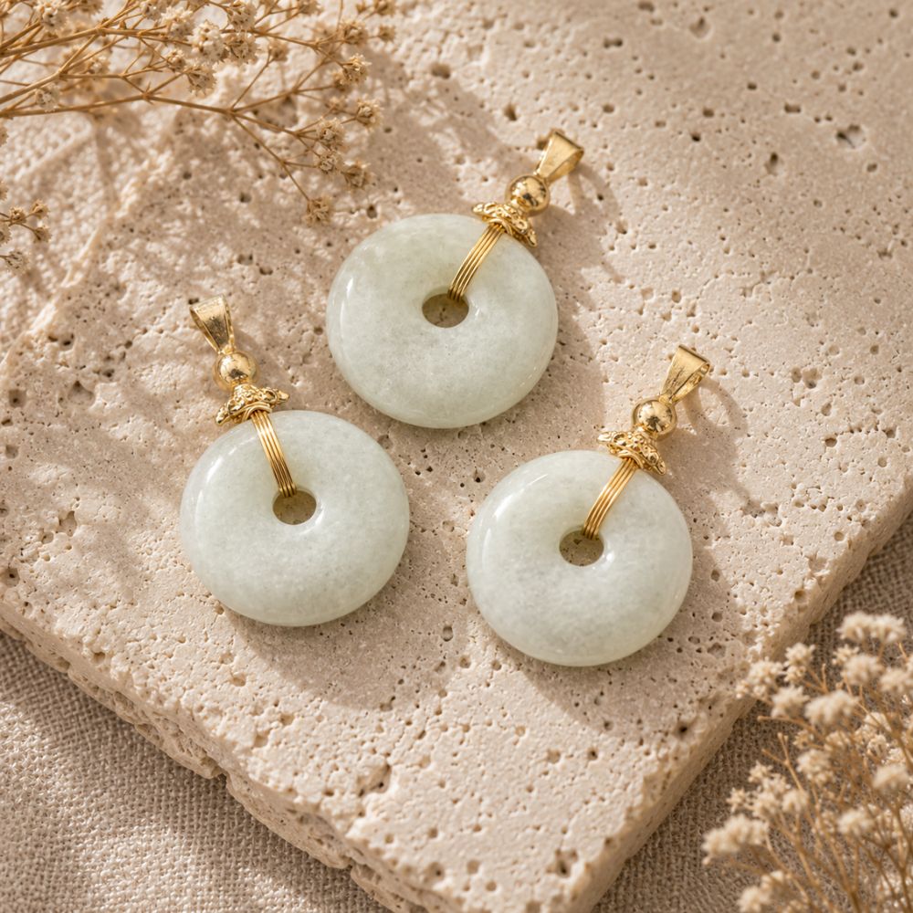 Jade Donut Pendant with natural green and white jade stone donuts arranged on a textured beige stone surface with soft linen fabric and dried flowers.