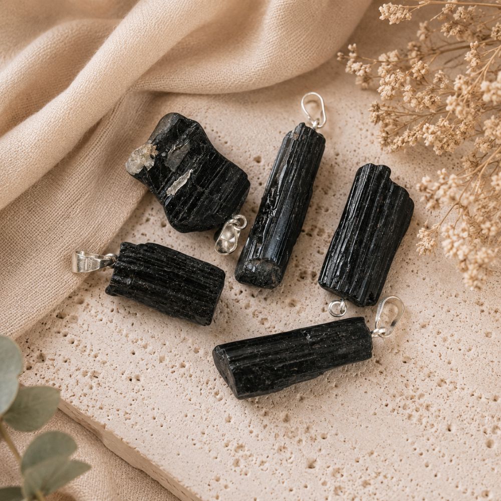 Five black tourmaline crystal pendants with silver jump rings, arranged on a textured beige stone tile with draped beige fabric and dried flowers.