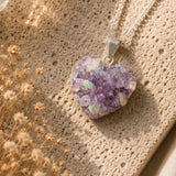 Heart-shaped purple druzy pendant on a silver chain against a textured beige background with dried flowers.