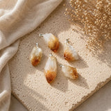 Four natural citrine crystal point pendants with silver bail loops, arranged on a textured beige stone slab with draped beige fabric and dried flowers.
