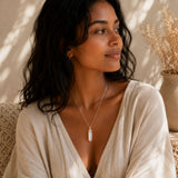 A woman with dark hair wearing a silver-toned crystal pendant necklace and a cream-colored wrap top, sitting in a warm, sunlit room with dried plants in the background.