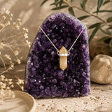 A gold-toned crystal pendant necklace displayed on a purple amethyst geode, set against a warm neutral background with dried flowers and stones.
