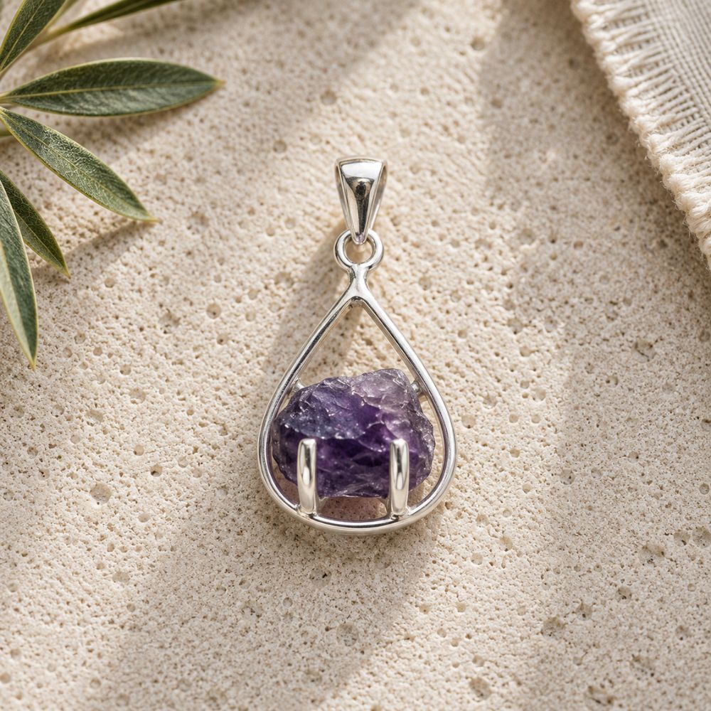 Raw amethyst teardrop pendant in a polished silver setting displayed on a natural stone surface with soft daylight and botanical accents.