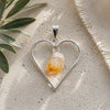 Raw citrine heart pendant featuring a natural citrine crystal suspended inside an open silver heart frame, photographed on a neutral travertine surface in soft natural light.