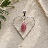 Raw pink tourmaline heart pendant featuring a natural pink tourmaline crystal suspended inside an open silver heart frame, photographed on a neutral travertine surface in soft natural light.