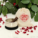 Meditation set with metal bowl, wooden stick, and drawstring bag with rose emblem on a white surface with red flowers and green leaves.