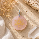 A round pink stone pendant with a gold-toned geometric symbol, hanging from a silver-toned bail, styled on a textured beige surface with dried flowers and a crystal cluster.