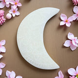 Decorative white crescent moon selenite charging plate on a brown background with pink flowers.