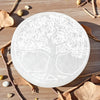 Round white selenite plate with tree of life engraving on a wooden surface with leaves and stones.