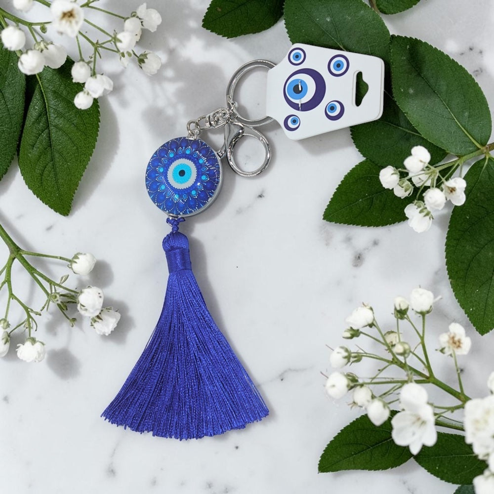 Blue evil eye keychain with tassel on a marble surface with white flowers and green leaves.