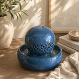 A blue ceramic water fountain with a perforated sphere and shallow round basin, styled on a wooden surface with books, a vase, and a textured cloth in a sunlit interior.