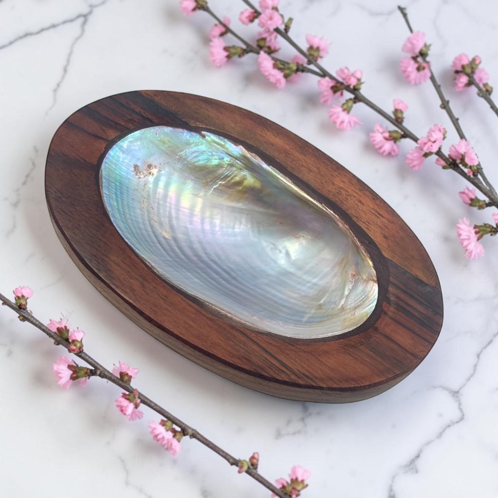 Wooden shell dish with a large shell on a marble surface with pink flowers.