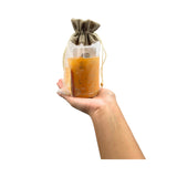Hand holding a glass jar with an orange substance inside, wrapped in a brown drawstring bag on a white background.
