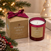 A holiday-themed scented candle in a dark red glass jar next to its kraft paper packaging box, both labeled 'Holiday Spice'. The scene is decorated with pine branches, red berries, and warm string lights on a marble surface.