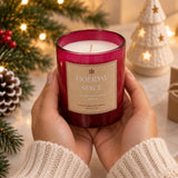 A person holds a hand-poured soy candle in a deep burgundy glass jar with a beige label reading 'Holiday Spice'. The background is a cozy, festive Christmas scene with warm lights, pine branches, and a small lit tree.