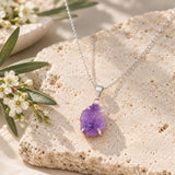 Raw purple amethyst crystal pendant in a silver prong setting on a delicate silver chain, displayed on natural beige stone with white flowers and greenery.