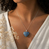 Person wearing a necklace with a blue heart-shaped pendant, surrounded by light-colored flowers.