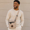 Man wearing a beige sweater and carrying a matching crossbody bag against a beige wall.
