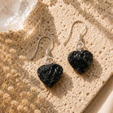 Heart-shaped black stone earrings on a textured beige surface with dried flowers.
