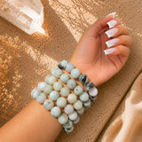 A close-up of a person's wrist wearing multiple beaded Jade bracelets made of pale green, white, and gray marbled stone, photographed on a textured tan surface with warm sunlight and shadows.
