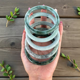 Hand holding a stack of green jade bracelets on a wooden surface with green leaves.