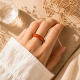 A close-up of a hand wearing a smooth, rounded Carnelianring, resting on a textured beige surface with dried flowers and warm sunlight.