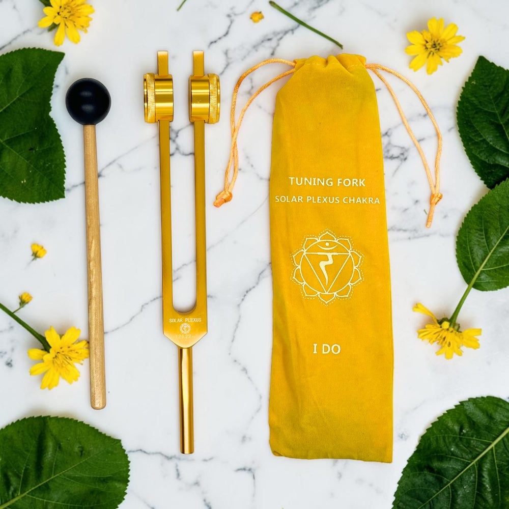 Gold tuning fork with a yellow pouch labeled 'Tuning Fork Solar Plexus Chakra' on a marble surface with leaves and flowers.