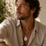 A man wearing a light beige linen shirt and a black cord necklace with a teardrop-shaped pendant featuring a white and yellow sun design, set against a warm, softly blurred background.