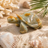 A polished tortoise figurine made of soapstone in muted green and amber tones, displayed on sand with seashells and coral.