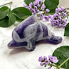 Purple and gray stone dolphin crystal on a marble surface with purple flowers and green leaves.