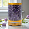A cylindrical crystal diffuser with purple stones on a reflective surface with tulips in the background.