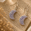 Crescent moon-shaped earrings with glittery finish on a textured beige surface with dried flowers.