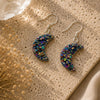 Colorful moon-shaped earrings on a textured beige surface with dried flowers.