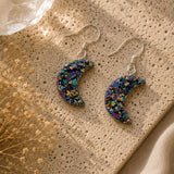 Colorful moon-shaped earrings on a textured beige surface with dried flowers.