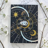 Decorative journal with celestial design on a marble surface with white flowers and branches.
