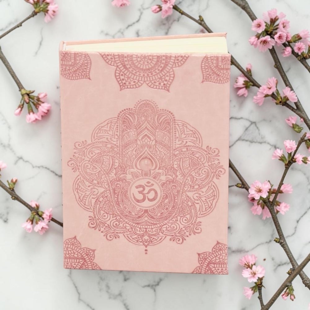 Earths Elements Pink Hamsa Journal - Small on a marble surface with cherry blossom branches.