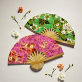 Two decorative fans, one pink with floral patterns and the other green with leaf designs, on a light surface with flowers around.